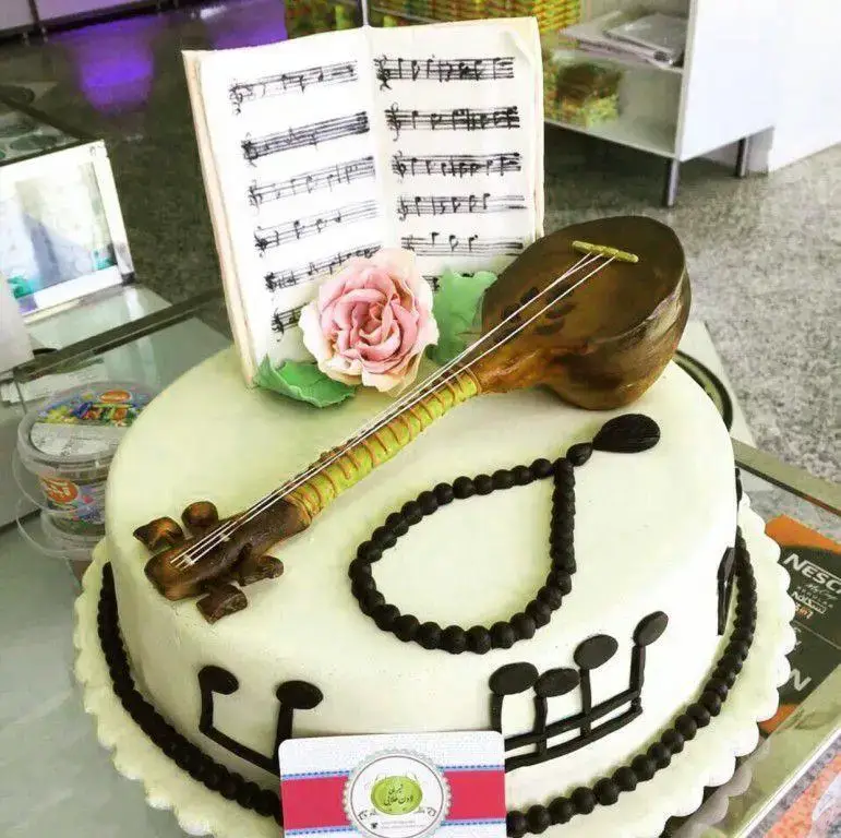 کیک فوندانت سفید با تزئینات مینیاتوری تار، نت‌های موسیقی و گل رز صورتی، اثری زیبا از شیرینی لادن طلایی برای مناسبت‌های خاص.