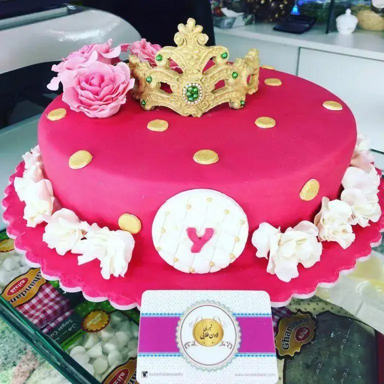 سفارش کیک تولد دخترانه صورتی با تم پرنسسی و تاج طلایی، گل‌های شکری و تزئینات شیک از قنادی لادن طلایی.