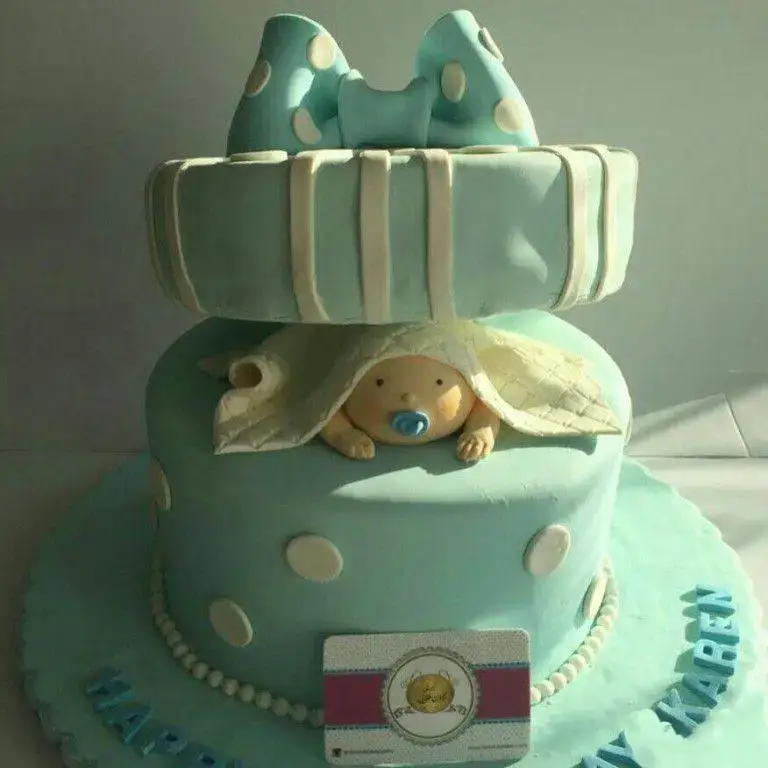 کیک جشن تولد یا تعیین جنسیت نوزاد، دو طبقه آبی روشن، با پاپیون بزرگ خالدار و عروسک نوزادی که از زیر پتو بیرون آمده، سفارش شیرینی لادن طلایی.