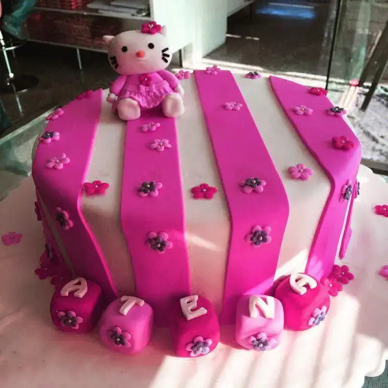 کیک تولد دخترانه فوندانت با تم هلن کیتی، راه راه صورتی و سفید با گل‌های زیبا و نام آتنا. شیرینی لادن طلایی.