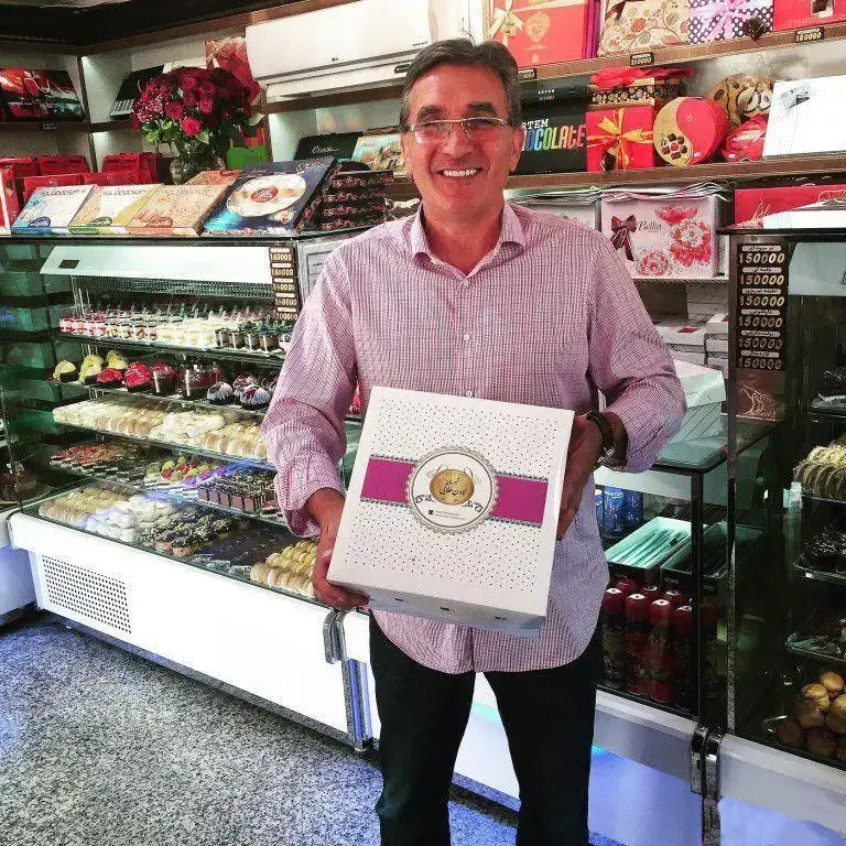 مردی خوشحال و خندان با جعبه شیرینی شیک لادن طلایی در قنادی. ویترین‌های پربار شیرینی‌های تازه و خوشمزه و بسته‌های کادویی زیبا.