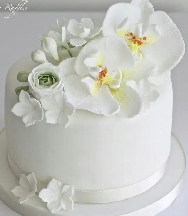 کیک عروسی مجلل سفید با گل‌های شکری ارکیده و رز سبز ملایم، اثری از شیرینی لادن طلایی، ایده‌آل برای جشن‌ها و مراسم خاص.