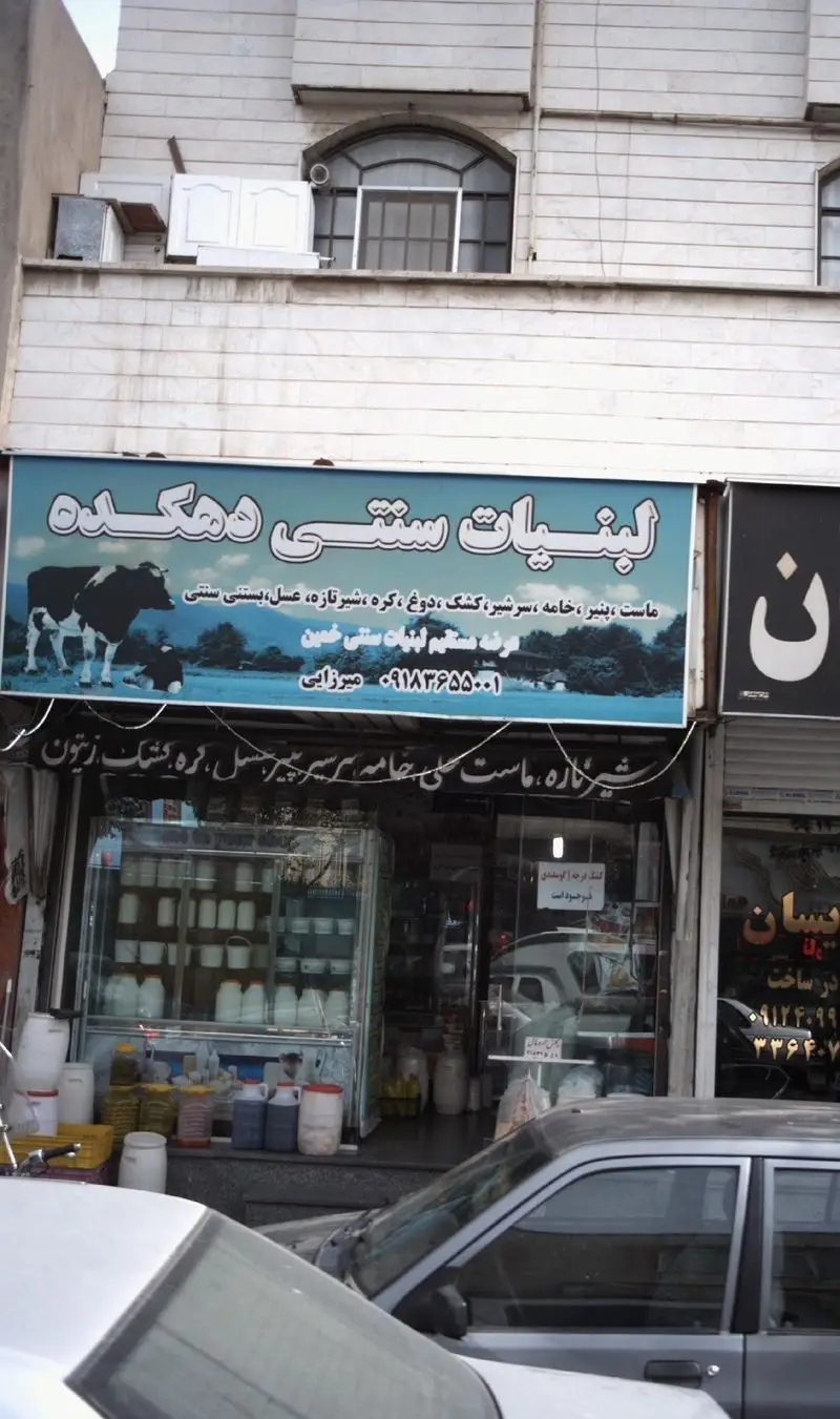 فروشگاه لبنیات سنتی دهکده با تابلوی حاوی گاو و منظره روستایی، عرضه‌کننده ماست، پنیر، شیر، دوغ، کره و عسل طبیعی.