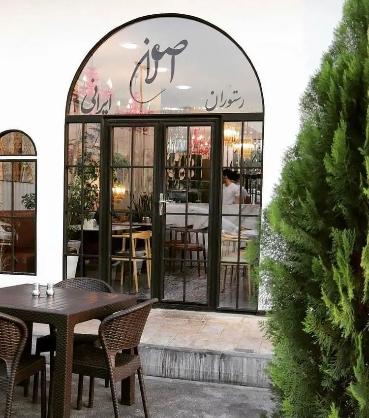نمای بیرونی رستوران اصلان، شامل ورودی شیشه‌ای با نام "اصلان" و "رستوران ایرانی" روی طاق پنجره. فضای باز با میز و صندلی.