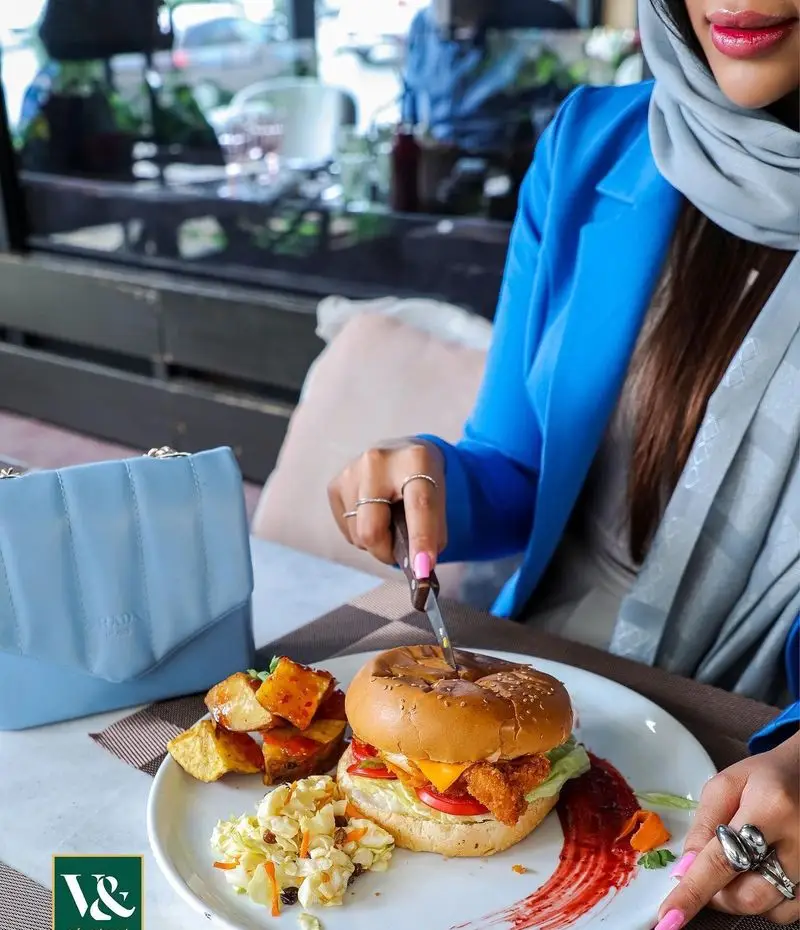 تجربه غذایی عالی در رستوران ایتالیایی ویند: خانمی در حال برش یک برگر لذیذ با سیب‌زمینی و سالاد تازه در محیطی دلنشین و شیک.