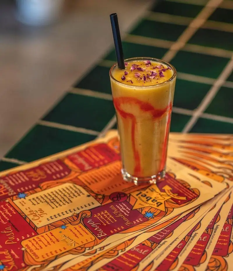 اسموتی میوه‌ای تازه و دلچسب کافه رستوران بابل با نی مشکی و تزئین گلبرگ رز، روی منوهای رنگارنگ و طرح‌های هنری خاص.