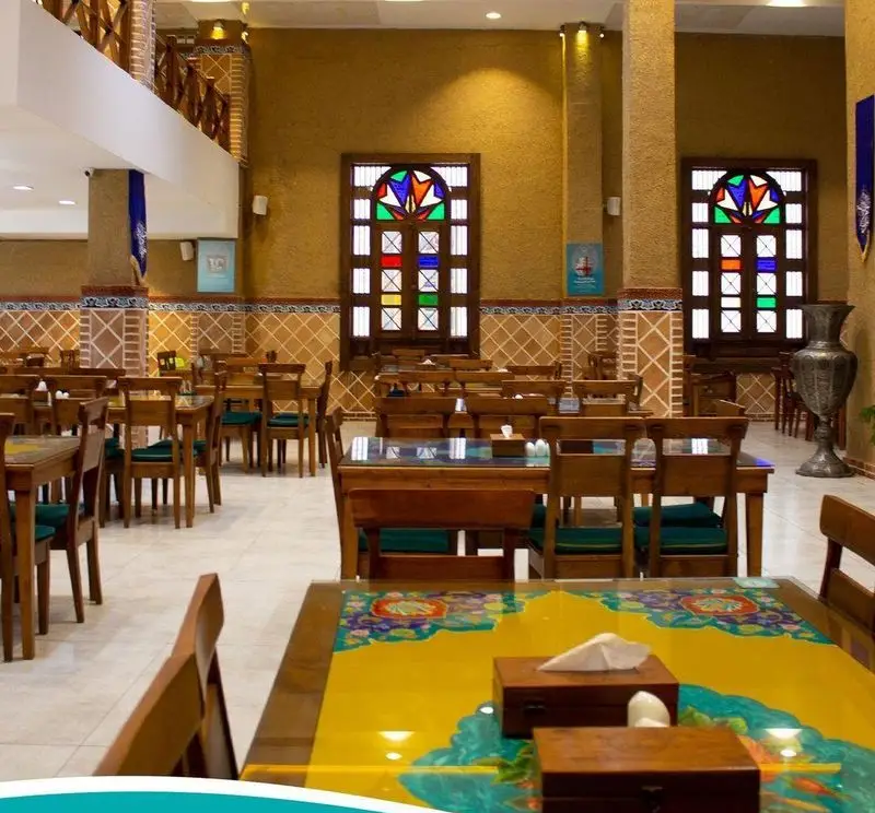 فضای داخلی رستوران سنتی مرشد سعادت آباد با دکوراسیون زیبا، میز و صندلی‌های چوبی و پنجره‌های رنگی در محیطی دلنشین برای پذیرایی.