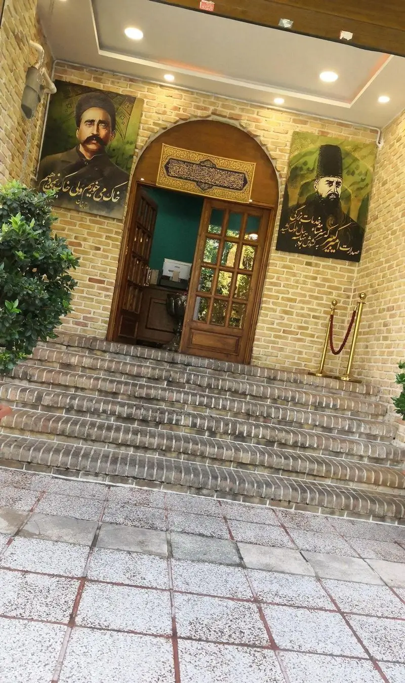 ورودی جذاب رستوران مرشد سعادت آباد با نمای آجری، پله‌های عریض، درب چوبی سنتی و پرتره‌های بزرگ تاریخی.