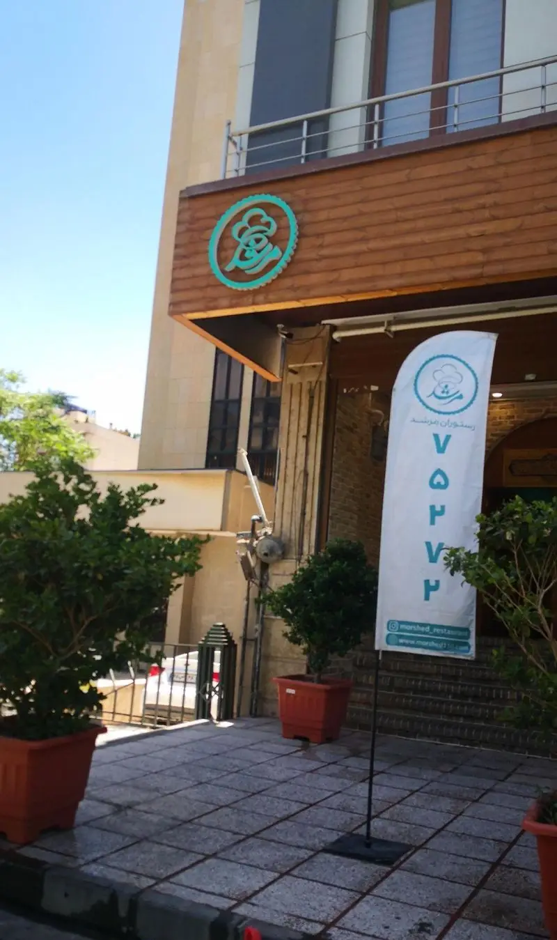 نمای بیرونی رستوران مرشد سعادت آباد با تابلوی اصلی و پرچم تبلیغاتی شامل لوگو، شماره تماس و آدرس وبسایت.