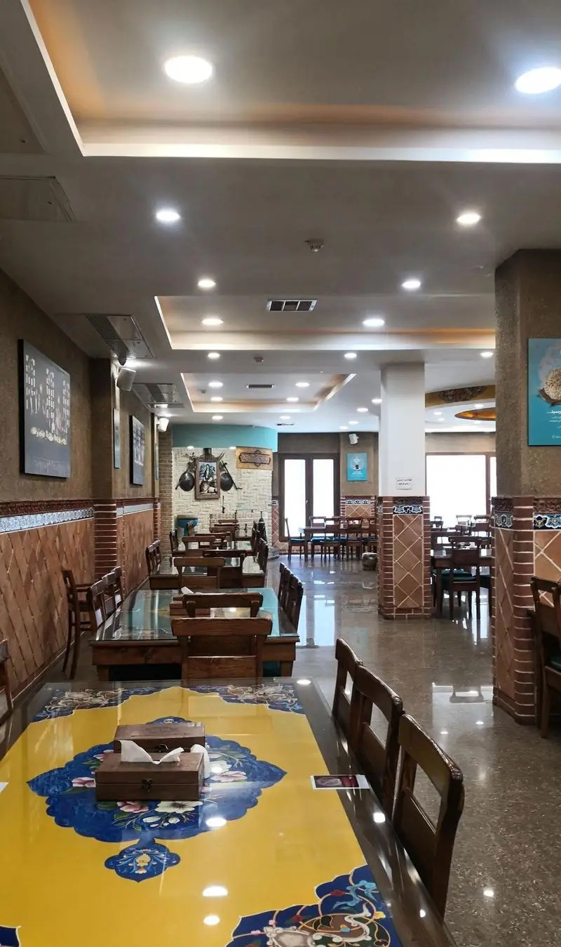 فضای داخلی بزرگ و روشن رستوران مرشد سعادت آباد با میزهای غذاخوری چوبی، رومیزی‌های سنتی آبی و زرد و دکوراسیون دیواری جذاب.