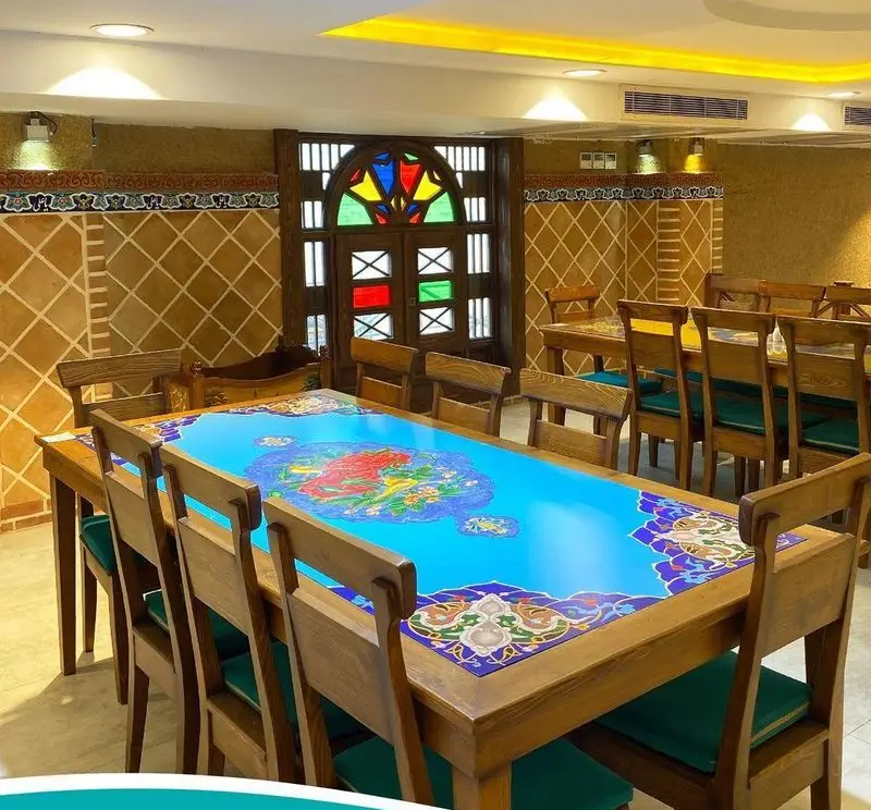 فضای داخلی زیبا و سنتی رستوران مرشد سعادت آباد با میز چوبی بلند و طرح آبی اصیل ایرانی، محیطی دلنشین برای پذیرایی.