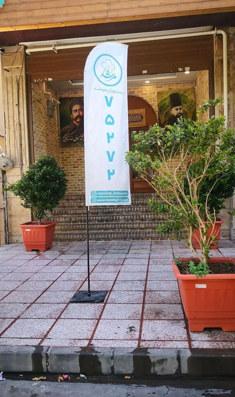 ورودی رستوران مرشد سعادت آباد، پرچم تبلیغاتی با لوگوی سرآشپز، شماره تماس ۷۵۲۲۵۰۷، آدرس اینستاگرام و وبسایت، گلدان‌ها و دیوارهای آجری.