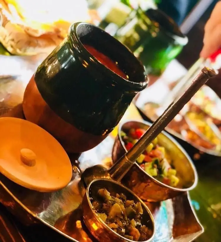 نمایی نزدیک از سس و مخلفات خوشمزه رستوران رافی، شامل سس قرمز در ظرف سفالی و ترشی و سالاد در کاسه‌های مسی شیک.