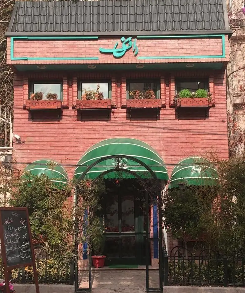 نمای بیرونی رستوران رافی با آجر قرمز، سقف تیره، سایبان‌های سبز ورودی و گلدان‌های گل. فضایی آرامش‌بخش برای لذت‌بردن از غذای ایرانی.