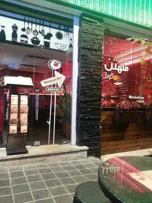 نمای بیرونی شیک رستوران منهتن گریل با ورودی شیشه‌ای، تزئینات جذاب و فضای نشیمن بیرونی، آماده پذیرایی از مشتریان.