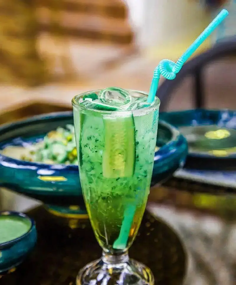 نوشیدنی خنک و جذاب سبز رنگ رستوران رزفر با یخ و برش خیار، سرو شده در لیوان شیک همراه نی فانتزی برای روزهای تابستانی.