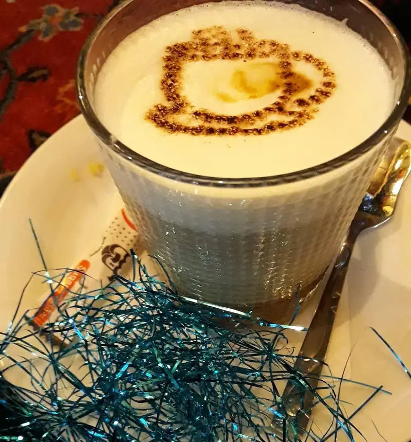 یک لیوان لاته آرت زیبا با طرح قلب کاکائویی، به همراه قاشق و تزیینات آبی در کافه رستوران شنزار. نوشیدنی گرم خاص و محیط دلنشین.