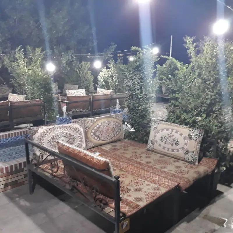 تصویری از فضای باز و زیبای رستوران سنتی شنزار در شب، با تخت‌های راحت و کوسن‌های طرح‌دار در میان فضای سبز دلنشین.