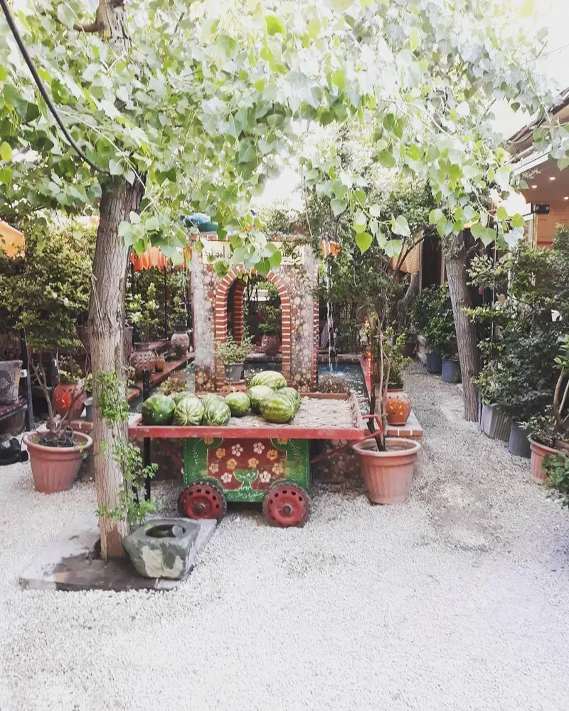 تصویری زیبا از فضای سرسبز و باغ‌ رستوران سنتی شنزار، با درختان، آبنمای سنگی و گاری تزئینی هندوانه‌ها.