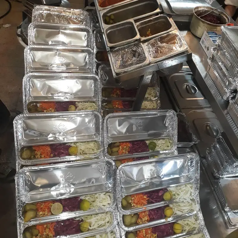 سینی‌های آلومینیومی پر از غذای آماده و دورچین‌های رنگی شامل ترشی، هویج، کلم و لیمو، از کترینگ رستوران شنزار.