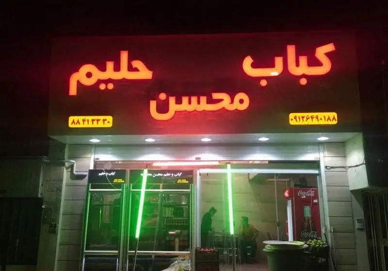 تابلوی درخشان کباب و حلیم محسن در شب با نئون قرمز و نورپردازی سبز، همراه با شماره تماس‌ها، یک رستوران فعال ایرانی.
