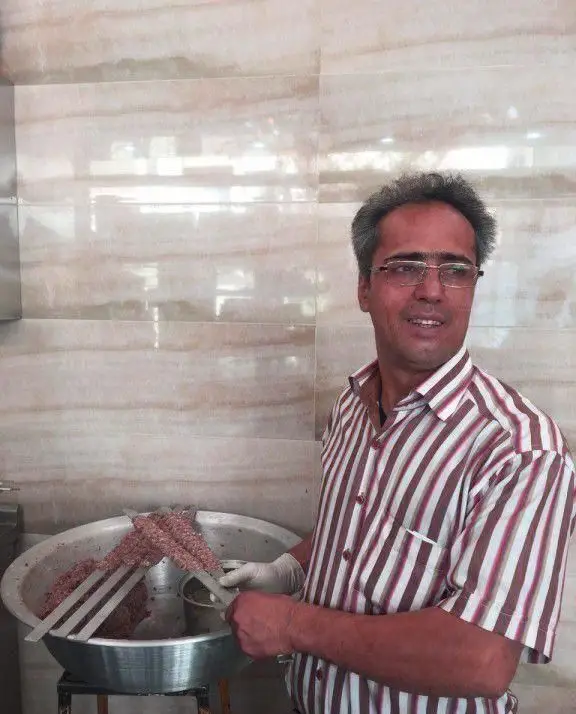 کباب سرای محسن: آشپزی ماهر با پیراهن راه راه و دستکش در حال سیخ کشیدن کباب کوبیده تازه، برای طبخ و سرو در کباب‌سرا.