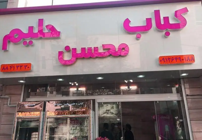 تابلوی صورتی رنگ کباب سرای محسن و حلیم، نمای خارجی مغازه با فونت برجسته، شامل شماره تماس‌ها و ورودی رستوران، مناسب برای سفارش غذای ایرانی.