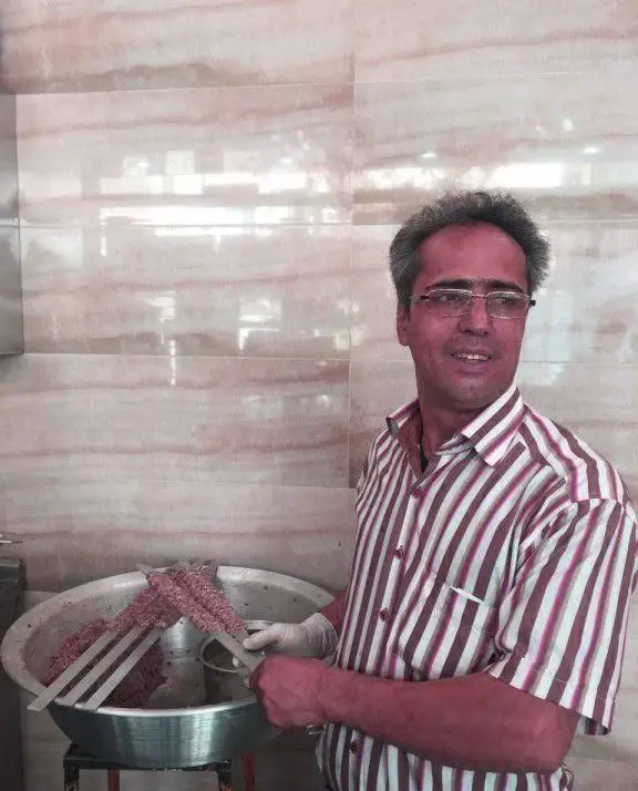 مردی لبخند بر لب، مشغول آماده‌سازی سیخ‌های کباب کوبیده تازه برای پخت در کباب سرای محسن، با رعایت بهداشت و کیفیت.