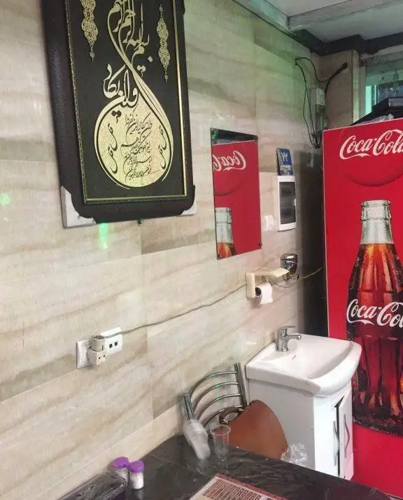 نمایی از فضای داخلی کباب سرای محسن با دیوارپوش سرامیکی، تابلوی خوشنویسی، سینک و یخچال بزرگ کوکاکولا، نشان دهنده محیطی کاربردی و مشتری‌پسند