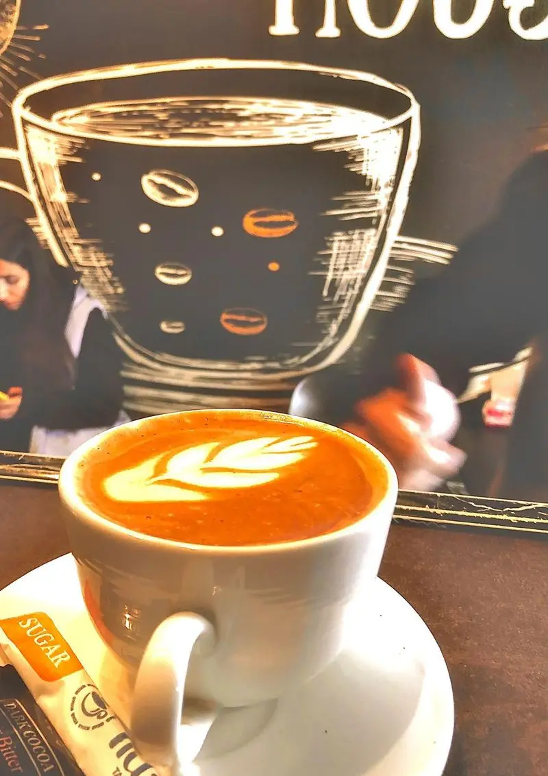 قهوه لاته گرم با هنر لاته برگ، کنار ساشه شکر در کافه علی. محیطی دنج با طراحی دیواری جذاب از فنجان قهوه و دانه ها.