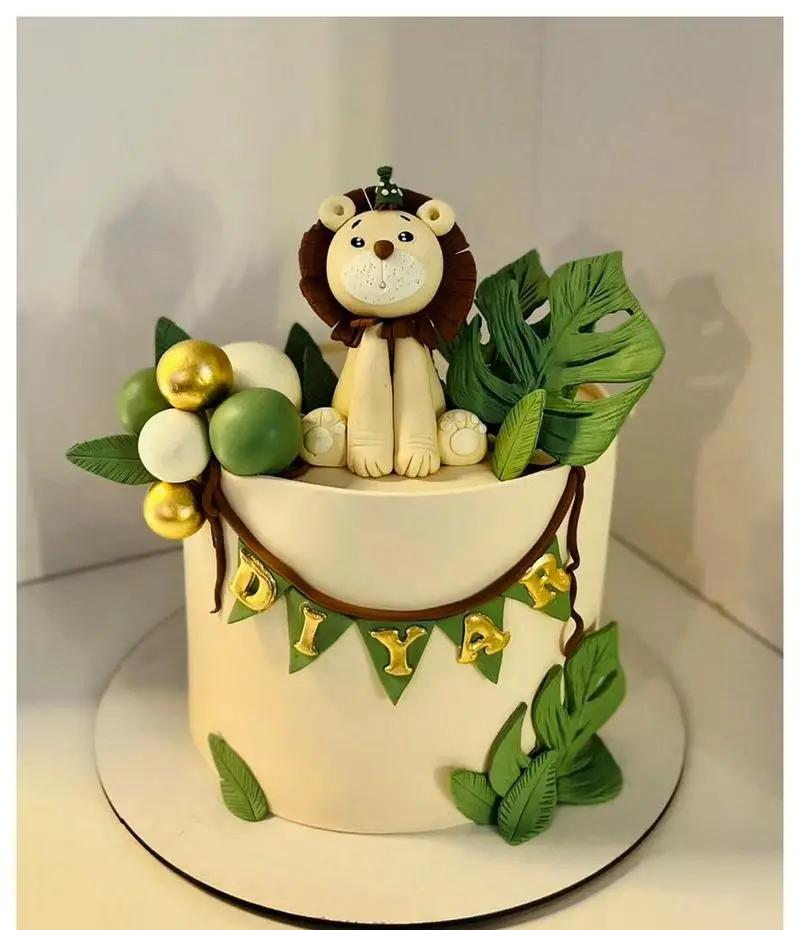 کیک تولد فوندانت با طرح شیر و جنگل، تزئین شده با برگ‌های سبز و توپ‌های طلایی و سبز. کیک خاص با اسم دیا از شیرینی مانلی تهرانپارس.