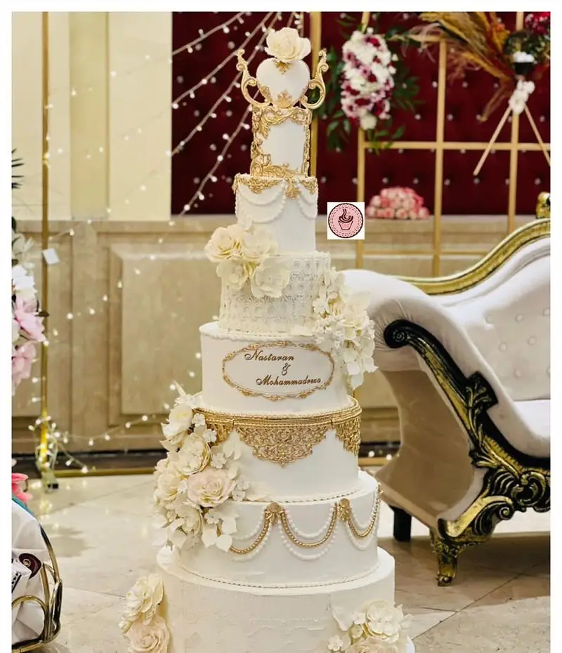 کیک عروسی لوکس و خاص از شیرینی مانلی تهرانپارس، کیک چند طبقه سفید با تزیینات طلایی، گل‌های شکری و نام عروس و داماد.