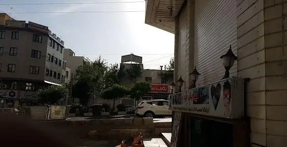 نمای بیرونی چشم‌انداز خیابانی کسب و کار فست فود "بمب جنت آباد" در طول روز با ساختمان‌های مجاور، فضای سبز و خودروهای عبوری.