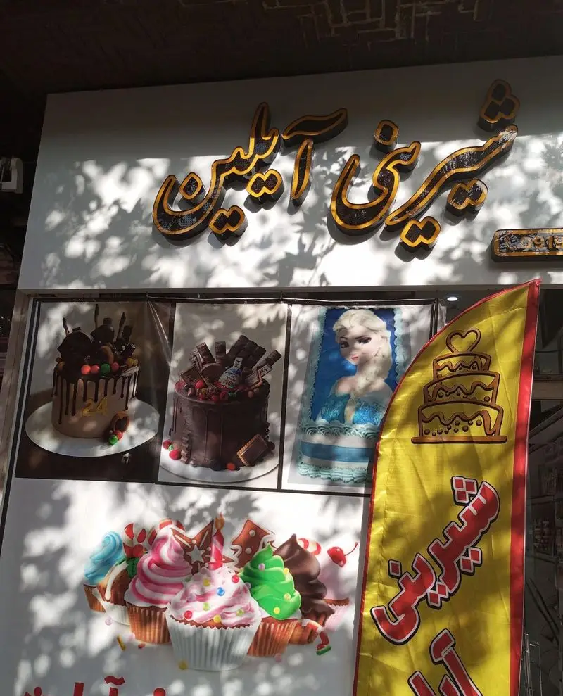 نمای خارجی قنادی شیرینی آیلین با تابلوی اصلی، بنر زرد رنگ و پوسترهای کیک‌های تولد، شکلاتی و کاپ‌کیک‌های متنوع و زیبا