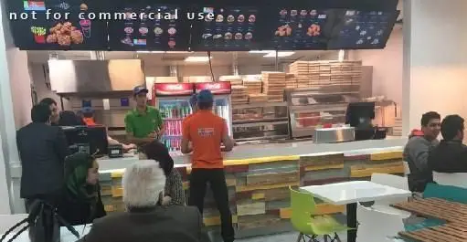 نمای داخلی فست فود سیتادو با مشتریان نشسته، پرسنل در حال خدمت و منوهای دیجیتال بالای پیشخوان، شامل پیتزا و مرغ سوخاری.