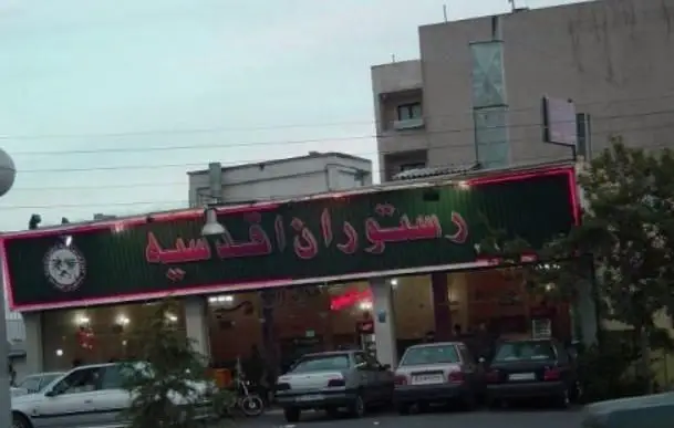 تصویری از نمای بیرونی رستوران ایرانی اقدسیه با تابلوی نورانی و روشن در شب، نمای خیابان و چند ماشین پارک شده در جلوی آن.