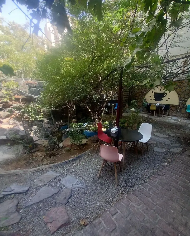 فضای سرسبز و دلنشین کافه پستو در فضای باز، با میز و صندلی‌های شیک و مدرن، مکانی ایده‌آل برای استراحت و نوشیدنی.