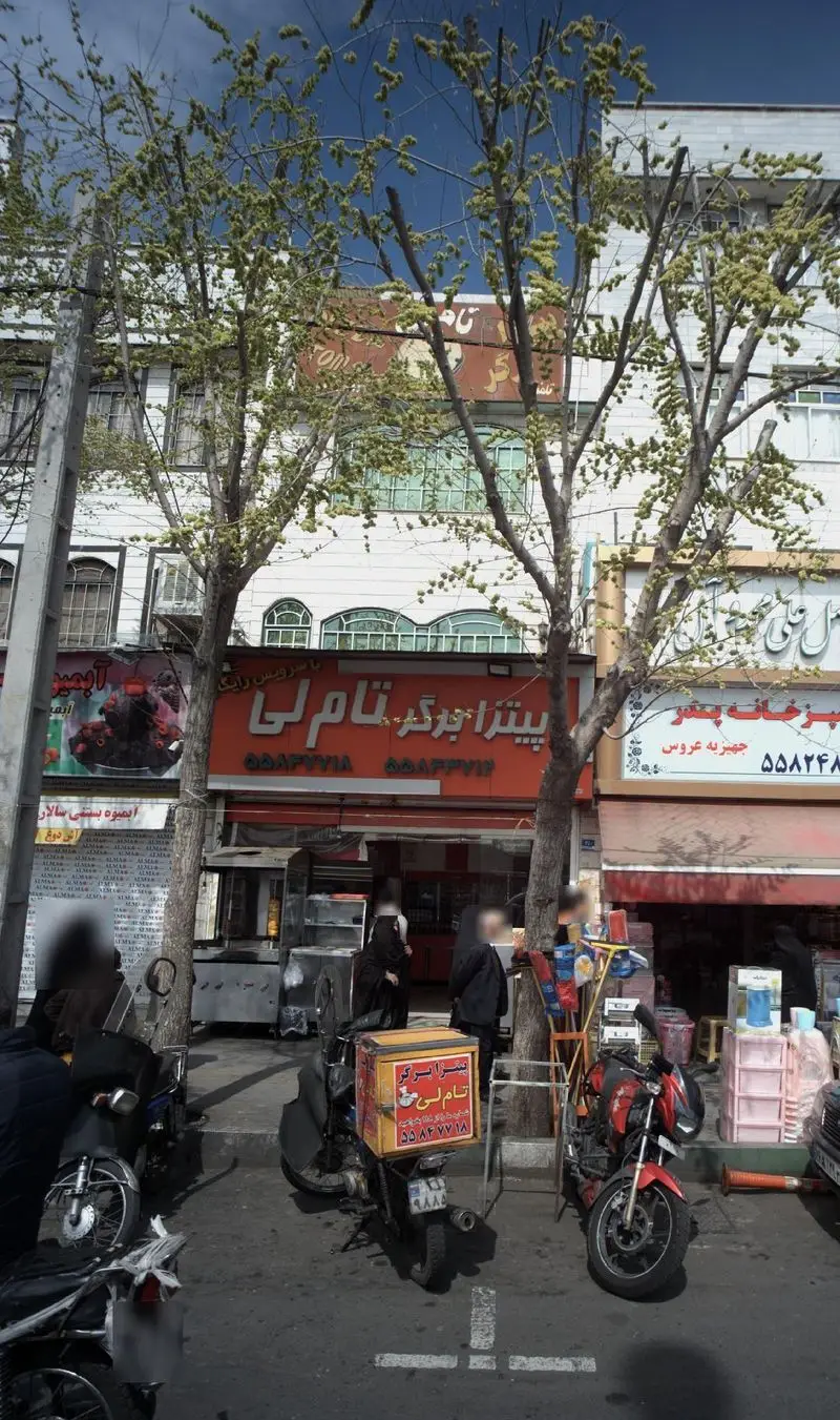 تصویری از نمای بیرونی پیتزا برگر تام لی واقع در یک خیابان شلوغ شهری در ایران، با موتورسیکلت‌های دلیوری و درختان سرسبز.