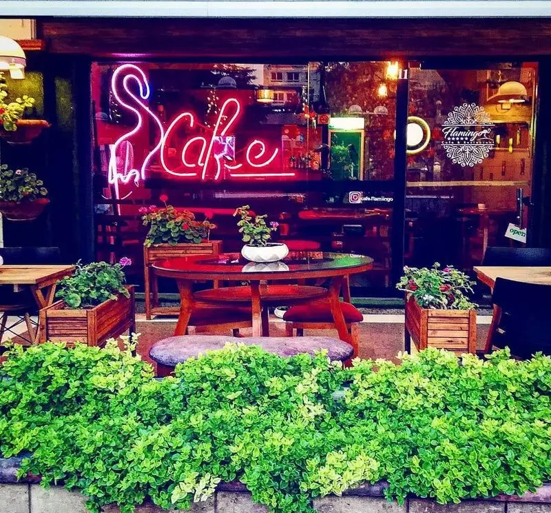 تصویر فضای بیرونی کافه شمعدونی با میز و صندلی‌های چوبی و گلدان‌های شمعدانی. نورپردازی گرم، محیطی دنج و آرام برای مشتریان.