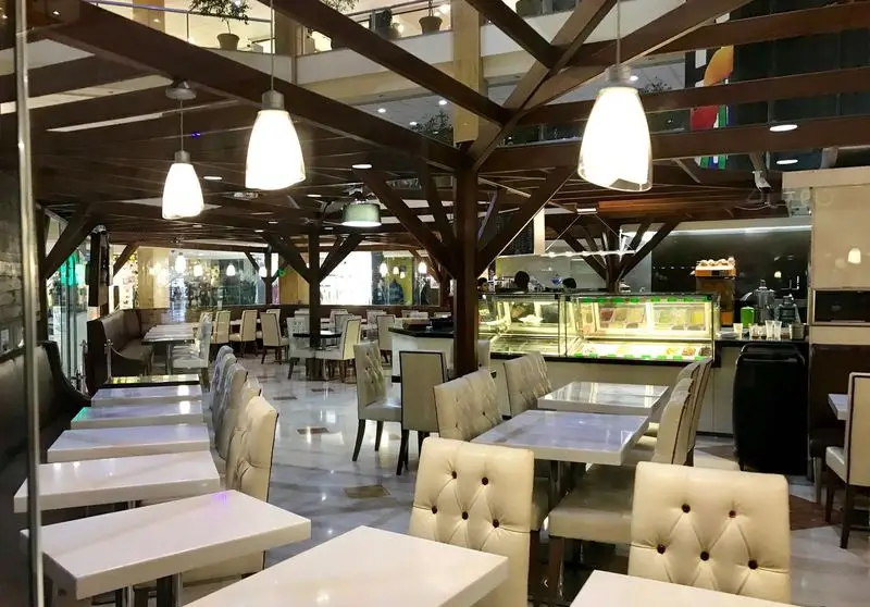 محیط داخلی لوکس و دلباز کافی شاپ گاندو با چیدمان میز و صندلی‌های سفید و راحت، سقف چوبی مدرن و ویترین جذاب دسرهای تازه.