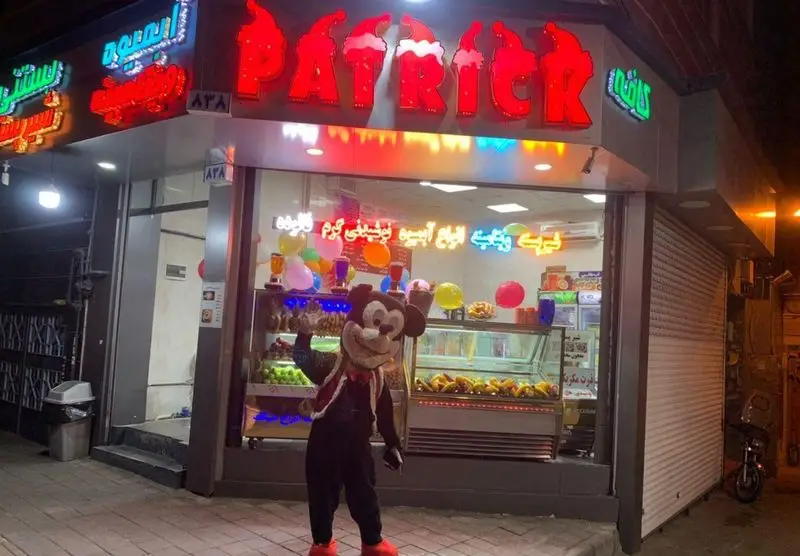 کافه آبمیوه و بستنی پاتریک، مغازه‌ای پرنور در شب با عروسک میکی‌ماوس. انواع آبمیوه، بستنی، فالوده و نوشیدنی‌های تازه ارائه می‌دهد.