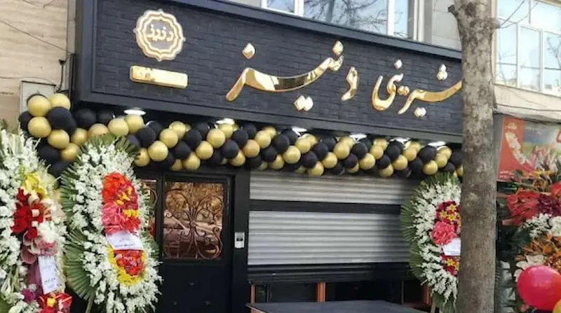 تصویر نمای بیرونی شیرینی فروشی دنیز در مراسم افتتاحیه با بادکنک‌های شیک مشکی و طلایی و تاج گل‌های تبریک.