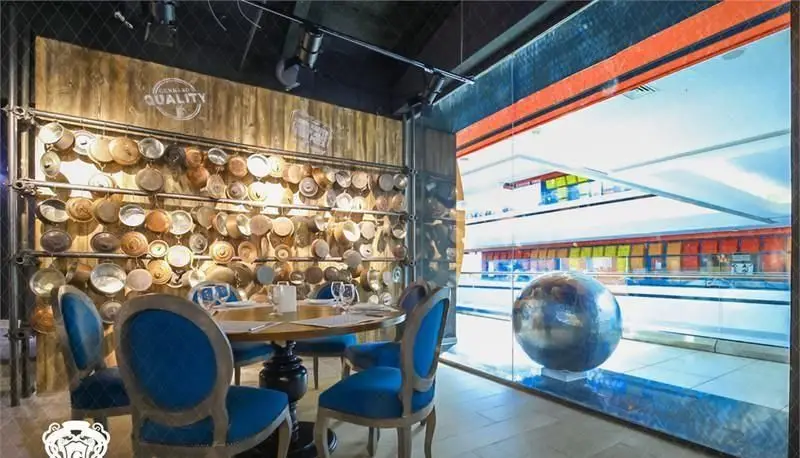 رستوران ایتالیایی جنارو کوروش: طراحی داخلی شیک با دیواری از ظروف فلزی قدیمی، میز ناهارخوری گرد و صندلی‌های سلطنتی آبی رنگ.