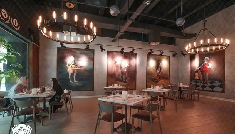 فضای داخلی رستوران ایتالیایی جنارو کوروش با دکوراسیون شیک، لوسترهای مدرن و نقاشی‌های بزرگ بر دیوار. میز و صندلی‌های چوبی برای مشتریان آماده است.