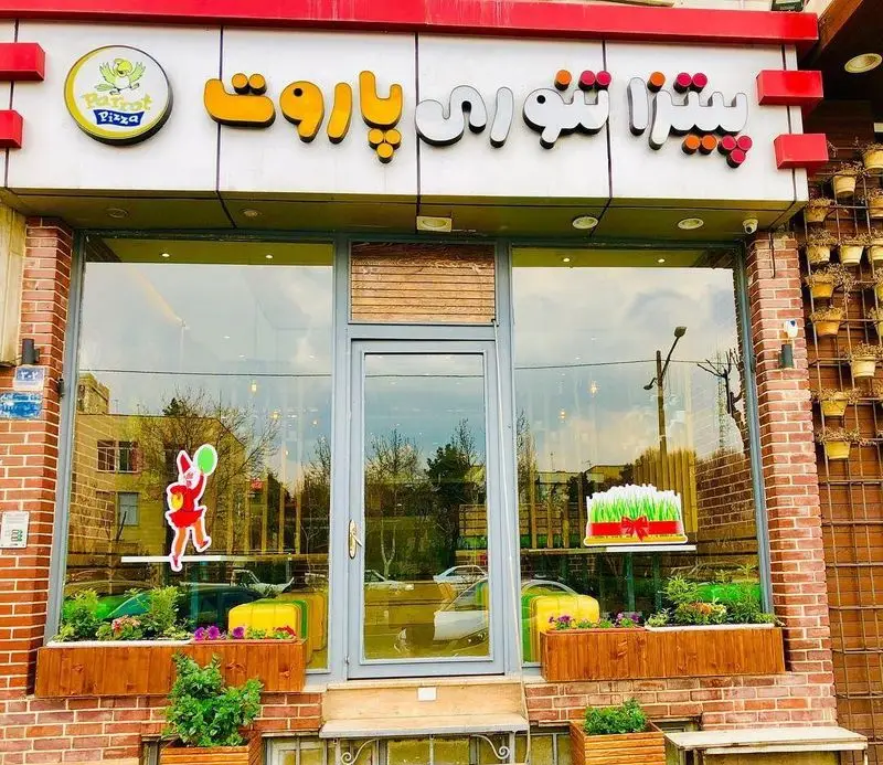 نمای بیرونی رستوران پیتزا تنوری پاروت با تابلوی روشن، لوگوی طوطی و ویترین شیشه‌ای زیبا شامل گلدان‌های گل و دکور نوروزی.