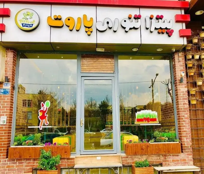 تصویر نمای بیرونی رستوران پیتزا تنوری پاروت. تابلوی روشن، ویترین شیشه‌ای و آرم پرنده، بهترین پیتزا و فست فود تنوری.