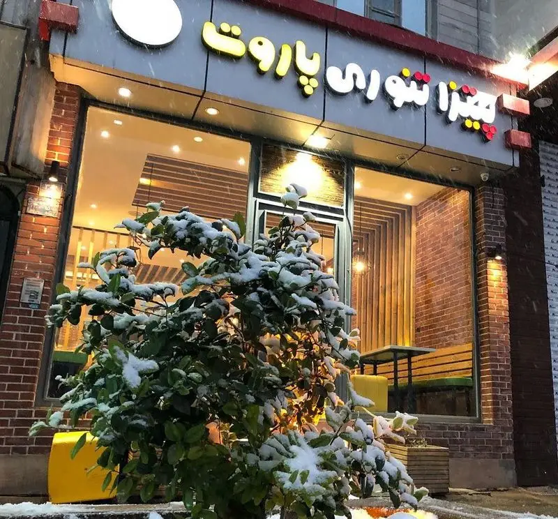 تصویر نمای بیرونی رستوران پیتزا تنوری پاروت در شبی زمستانی و برفی، با تابلوی روشن و درختچه پوشیده از برف، فضایی دلنشین.