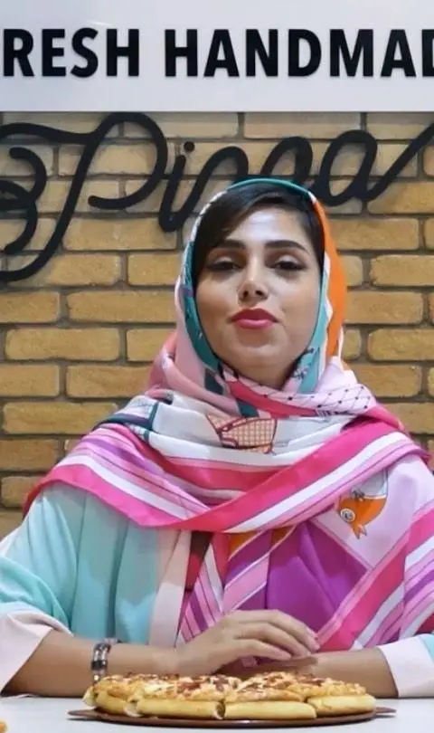 زن با روسری رنگی و شاداب در محیط تله پیتزا (شعبه پیروزی)، در کنار پیتزاهای تازه و دست‌ساز، آماده پذیرایی از مشتریان.