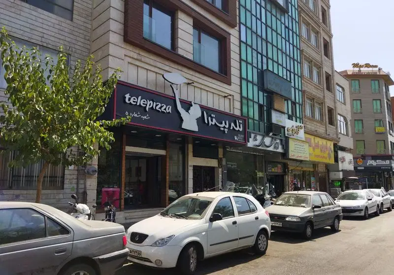 تصویری از نمای خارجی شعبه پیروزی تله پیتزا، فست فود و رستوران پیتزا در تهران با لوگوی Telepizza و خودروها در خیابان.