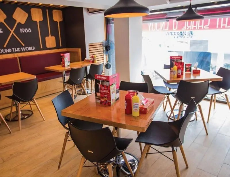 فضای داخلی مدرن رستوران تله پیتزا شعبه پیروزی با میز و صندلی های چوبی و مشکی، دکور دیواری پیتزا و محیطی دلنشین برای مشتریان.